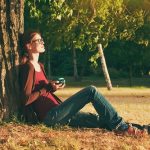 Smiling girl with cup of tea or coffee enjoying near park tree in morning sunlight